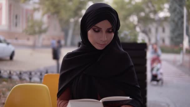 屋外のカフェに座っている間、黒ヒジャーブ読書本の女の子、試験の準備、イスラム教徒の女性の間での教育。正面図 — ストック動画