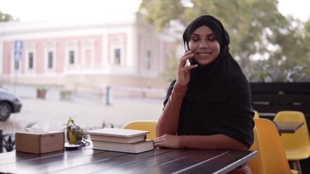 Genç, tesettürlü, tesettürlü, gülümseyen, kafede oturan, neşeli Müslüman bir kadın. Masada kitap bulundurmak. Arka planda bulanık, güzel bir bina. — Stok video
