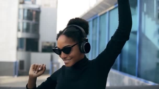 Close-up retrato de adorável afro menina feliz sorrindo. Mulher africana deslumbrante com pele castanho-clara relaxante em fones de ouvido e dança engraçada no fundo da cidade moderna. Vestindo camisola preta e — Vídeo de Stock