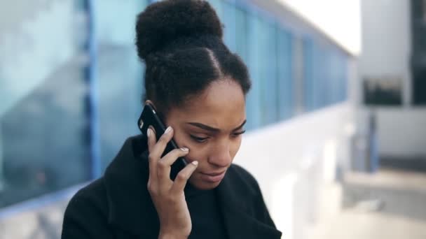 Крупный план портрета черной женщины, говорящей по мобильному телефону, решающей деловые вопросы удаленно. Африканская привлекательная женщина что-то обсуждает, серьезное лицо. Стоя рядом с офисным зданием — стоковое видео