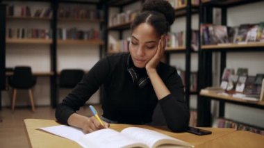 Çekici, konsantre olmuş Afrikalı genç bir kadın masanın başında oturuyor ve modern kütüphanedeki üniversite için not defterine bazı notlar yazıyor, sınavlara hazırlanıyor. Siyah kazak ve kulaklık takıyor.