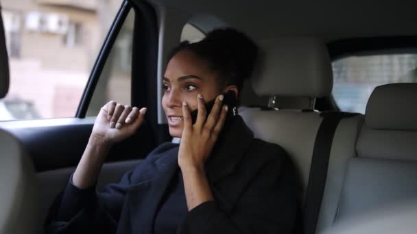 Hermosa mujer de negocios afroamericana sentada en el asiento trasero de un taxi con salón de cuero brillante, hablando por su teléfono móvil. Una mujer relajada hablando por teléfono en el coche. Movimiento lento — Vídeos de Stock