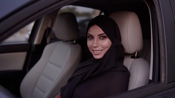 Mulher muçulmana jovem bonita em preto tradicional hijab em uma cabeça sentada no carro, no assento do motorista. Menina com uma maquiagem perfeita olhando para a câmera e sorrindo — Vídeo de Stock