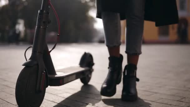 Close up de pernas de uma jovem mulher motorista de scooter andando com seu transporte ecológico, scooter elétrico. Menina andando com scooter preto vestindo jeans e botas elegantes. Luz solar no fundo — Vídeo de Stock