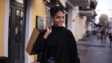 Küçük kahverengi bir çantayla sokakta yürürken gülümseyen çekici melez bir kızın portresi. Lüks sokaklardan alışveriş yapan genç ve mutlu bir kadın.
