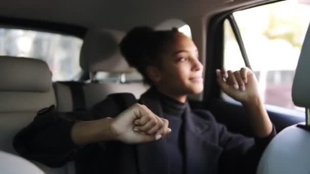 Mujer afro-americana despreocupada en ropa negra baila mientras está sentada en un asiento trasero, escuchando la radio o emocionada por el viaje. Mirando la ventana. Durante el día, paisaje urbano — Vídeo de stock