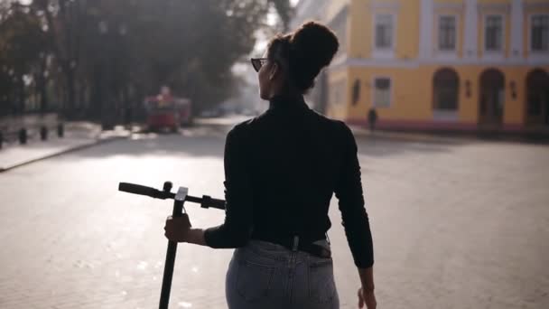 Vzácný pohled záběry mix závodil žena procházející po ulici města. Dívka v černém plášti a slunečních brýlích kráčí s elektrickým koloběžkou v ranním městě, ohlíží se přes rameno. Pomalé — Stock video