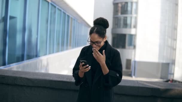 Mulher afro-americana tem mensagem surpresa, lê notícias impressionantes no telefone celular. Menina excitada começando a gesticular, a dançar ao ar livre, na rua. Vestindo casaco preto, jeans e óculos — Vídeo de Stock