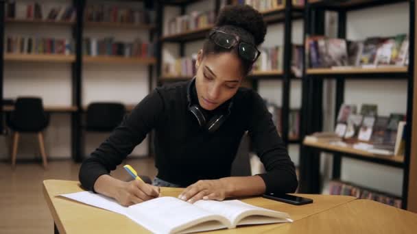 Jovem africana concentrada atraente sentada ao lado da mesa e fazendo algumas anotações em seu livro de anotações para a universidade na biblioteca moderna, preparando-se para exames. Vestindo camisola preta e fones de ouvido — Vídeo de Stock