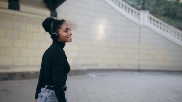 Chica afro alegre feliz sonriendo. Impresionante mujer africana con la piel de color marrón claro relajante en los auriculares y bailando lentamente en el fondo del edificio de la ciudad vieja. Usando suéter negro y jeans elegantes. Despacio. — Vídeo de stock