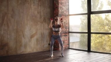 Gri spor üniformalı çekici bir kadın. Pencere arkasından güneş ışığı altında spor salonunda dambıllarıyla çömeliyor. Yavaş çekim