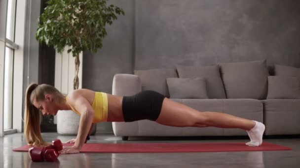 Zdrowa młoda kobieta w odzieży sportowej - żółta bluzka i czarne szorty robiące pompki na macie fitness przed szarą sofą w salonie. Młoda kobieta ćwiczy w domu. Widok boczny — Wideo stockowe