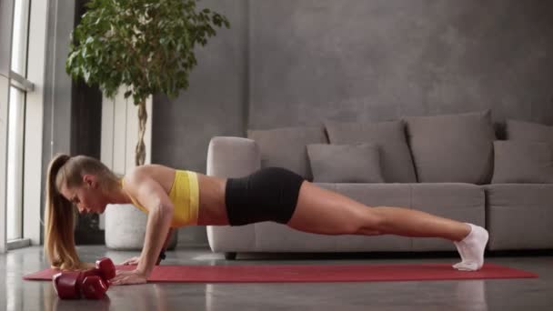 Mujer joven y saludable en ropa deportiva - top amarillo y pantalones cortos negros haciendo flexiones en la alfombra de fitness en frente del sofá gris en la sala de estar. Mujer joven haciendo ejercicio en casa. Vista lateral — Vídeo de stock