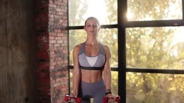 Boş fitness kulübünde küçük kırmızı halterli bayan sporcu ya da antrenör eğitimi. Genç bir kadın, modern spor salonlarında ağırlığı kullanarak el eğitimi veriyor. Spor salonundaki kadın kameranın önünde kollarını kaldırıyor.