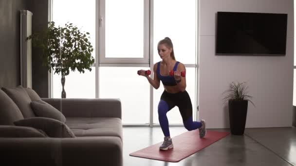 Stark fitness kvinna lyfta hantlar medan du gör lungor, njuta av ordentlig andning under träningen. Ganska atletisk kvinna gör sport motion hemma på en matta — Stockvideo