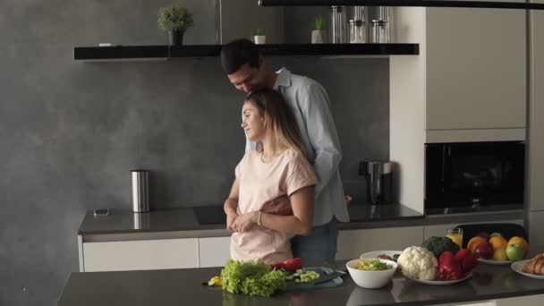 迷人的年轻的高加索夫妇一清早在家里现代化的厨房见面。 站在柜台前拥抱，然后妻子亲吻她的丈夫。 美满幸福的夫妻享受早晨 — 图库视频影像