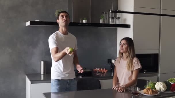 Attraktiv kærlig kaukasiske par have det sjovt i hjemmet køkkenet. Flot mand i jeans og hvid T-shirt jonglerer med æbler for at imponere sin kæreste, hun krammer ham. Langsom bevægelse – Stock-video