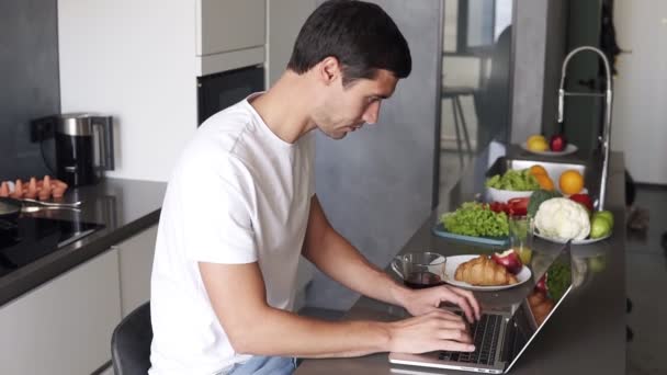 若い男が座って、キッチンカウンターで自宅で午前中にノートパソコンの仕事を使用しています。マグカップからお茶を飲みながら、メールやソーシャルメディアのインターネットをチェックする男 — ストック動画