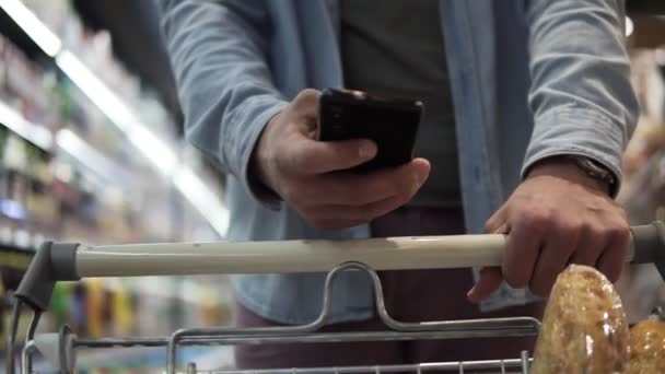 若い男性の手の映像を閉じ、スタイリッシュな時計が背景にぼやけた背景やスーパーマーケットでの買い物にトロリーカートを押しています。現代のスマートフォンを使って — ストック動画