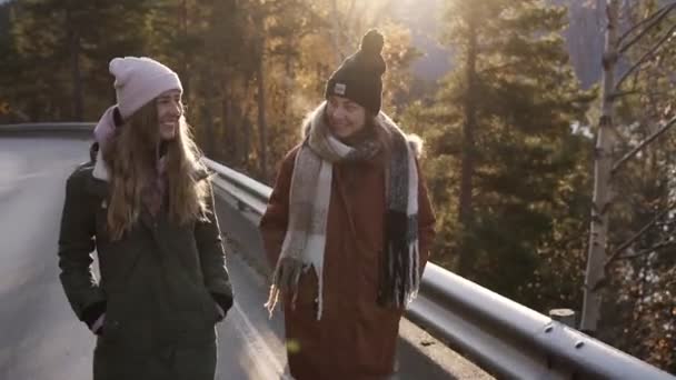 Mujeres turistas positivas en abrigos de invierno caminando en cámara lenta en un largo camino a través del campo hacia los picos nevados de las montañas, árboles dorados y el lago en el fondo. Sonriente caucásico — Vídeo de stock