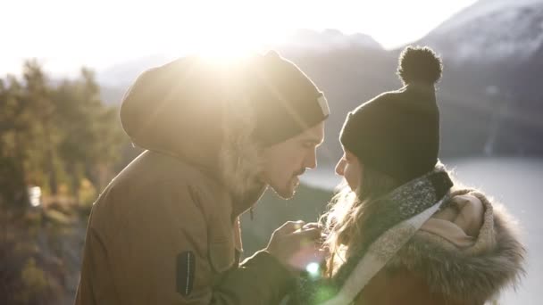 Joven pareja de pie abrazar besos, mirando paisaje soleado día de invierno. Mujer y hombre en ropa de abrigo de pie en frente hermoso paisaje de las montañas noruegas picos y lago de invierno. Bengalas de lente — Vídeo de stock