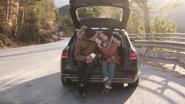 Een liefdevol koppel in een road trip. De man en het meisje zitten in de open kofferbak van de auto sandwiches te eten. Ze praten, kussen. Stop buiten in het midden van de weg — Stockvideo