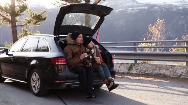 Una coppia amorevole in viaggio. Il ragazzo e la ragazza sono seduti nel bagagliaio aperto della macchina, a mangiare panini. Ammirando la vista intorno - bella natura della Norvegia. Fermati all'aperto in mezzo alla — Video Stock