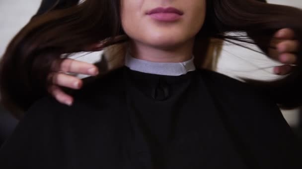 Young woman looking to the camera after haircut. Hairdresser waving clients long brown hair after haircut. Cropped footage of a gorgeous brunette girl in barbershop. Front view — Stock Video