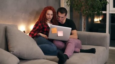 Gri dizüstü bilgisayarlı güzel bir çift kanepede oturuyor. Çift - kısa boylu bir adam ve kızıl saçlı kız dizüstü bilgisayarda bir şey izliyorlar akşam şık bir oturma odasında kanepede oturuyorlar.