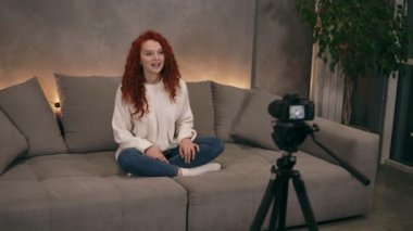 Çekici kıvırcık saçlı genç kız vlogger internet blogu için kamera önünde konuşuyor, gülümsüyor. Kadın kot pantolon ve beyaz kazak giyiyor.