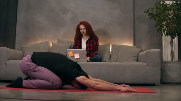 Joven pareja caucásica en la relajante sala de estar en casa, rizado firl readheaded utilizando un moderno portátil inalámbrico en un sofá gris mientras su novio está haciendo yoga, estirándose sobre una alfombra roja. Vista frontal. Loft. — Vídeo de stock