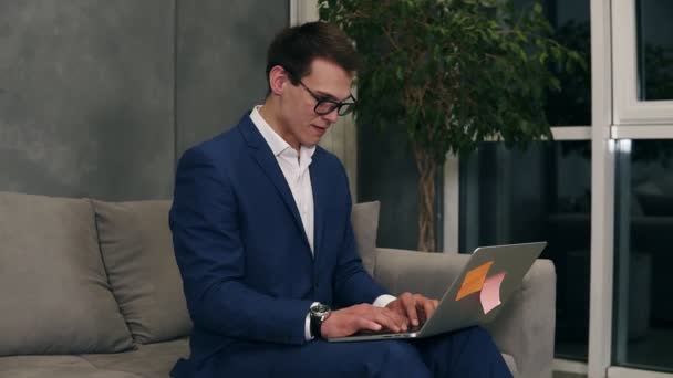 Hombre de negocios caucásico con traje azul formal y gafas elegantes sentado en el sofá escribiendo en su computadora portátil de rodillas en la habitación del hotel. Loft habitación interior — Vídeos de Stock