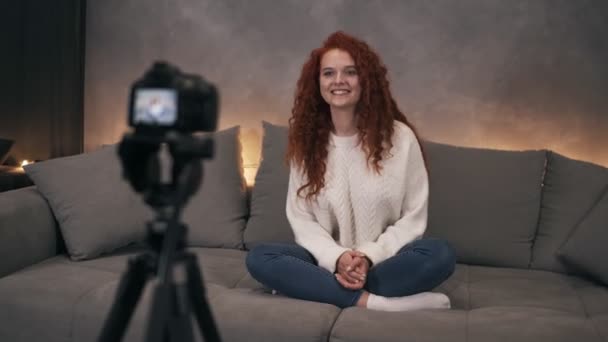 Atrakcyjne kręcone czerwono-głowy młoda dziewczyna vlogger mówi przed kamerą nagrywania wideo dla bloga online w Internecie mówienie, uśmiech i gestykulacji. Kobieta nosi dżinsy i biały sweter. — Wideo stockowe