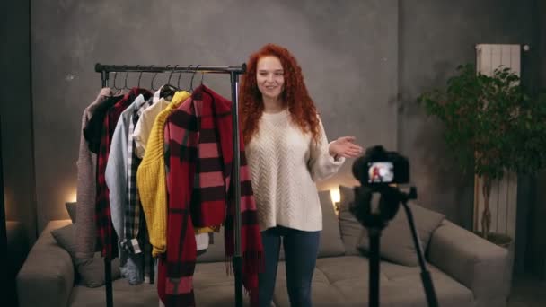 Elegante vlogger mulher jovem de cabeça vermelha está gravando vídeo para blog on-line sobre roupas da moda em pé nextto o rack com roupas coloridas falando e gesticulando. Vlogging, pessoas e conceito de casa — Vídeo de Stock