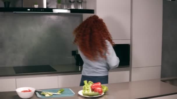 Happy active red headed young woman holding spoon as a microphone singing songs and dancing while cooking in kitchen, carefree girl preparing vegetable salad healthy food having fun alone at home — ストック動画