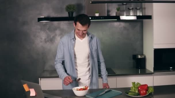 Joven súper emocionado sosteniendo cuchara como un micrófono cantando canciones y bailando mientras cocina en la cocina, hombre despreocupado preparando ensalada de verduras comida saludable divirtiéndose solo en casa. Se siente como un — Vídeos de Stock