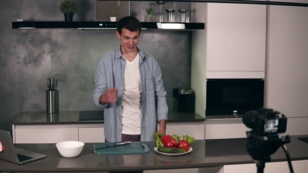 Young man in casual clothes leads a culinary blog and records video in grey kitchen. Video shooting of the blog - welcomes his followers and starting to cut vegetables on a dashboard. Modern marketing — ストック動画