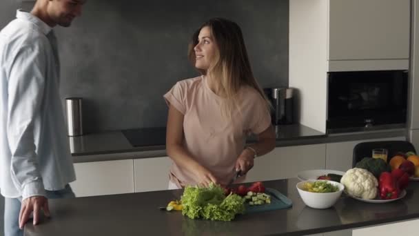 A young man is kissing his girlfriend when shes making a fresh salad for them. He came to hug her and appreciate. Long haired woman kisses him back. Modern kitchen in grey colours — Stock Video