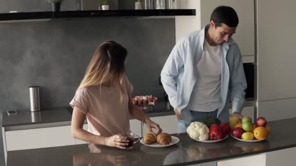 Morning at the kitchen counter, fast breakfast before work - croissants, juice and coffee for breakfast. Handsome husband came to the kitchen, welcomes his wife with a kiss, sipping an orange juice — Stock Video
