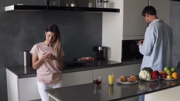 Young couple looking at something on their smartphones screen in the kitchen with modern loft design. Reading morning news or checking social media. Standing separately from each other — Stockvideo