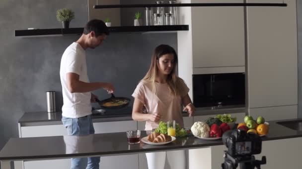 若いです魅力的なカップルブロガーでカジュアル服撮影ビデオフードブログ彼らの日常生活について,現代のキッチンでカメラで簡単な朝食を調理 — ストック動画