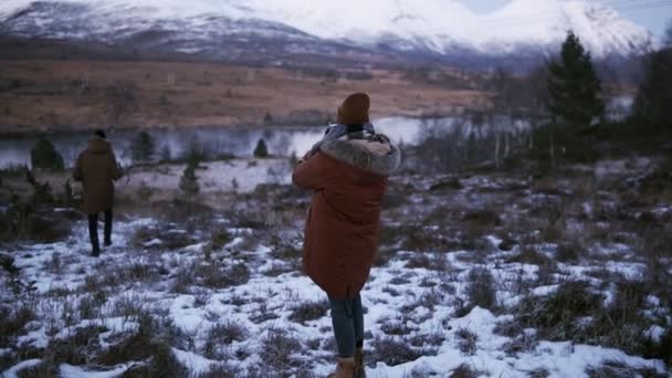 Two tourists walking in slow motion through the countryside towards the snowy mountain peaks on the background. A person on a winter coat trying to take a photo of beautiful Norway nature — Stockvideo