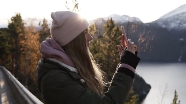 戴米色帽子的女人站在路上，用她的智能手机，向地平线望去，用她的智能手机，为美丽的挪威风景拍照，那里有高山雪峰和湖泊。 侧视图 — 图库视频影像