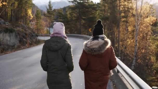 Due turisti di sesso femminile che camminano al rallentatore su una lunga strada attraverso la campagna verso le cime innevate delle montagne e gli alberi dorati sullo sfondo. Vista rara — Video Stock