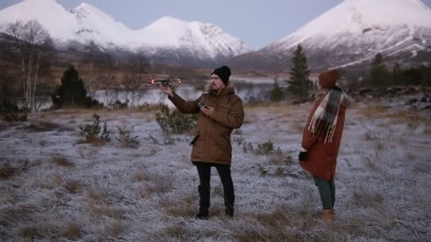 A caucasian couple launches drone at the field in front the mountains with snowy peaks on the background. Man operating the copter, woman standing close. Snowy countryside around — Stock Video