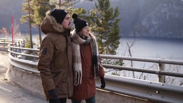 Turistas en abrigos de invierno y sombreros negros caminando en cámara lenta en un largo camino a través del campo hacia los picos nevados de las montañas y el lago en el fondo. Sonriente pareja caucásica explorando — Vídeos de Stock