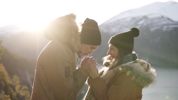 Молодая пара в зимних пальто стоят друг перед другом, держа за руки великолепные снежные горы и озеро, мужчина согревает руки своих подружек и целует их. Концепция путешествия. Солнце сильно светит — стоковое видео