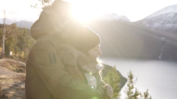 Jovem casal feliz em casacos de inverno abraçando, as pessoas abraçando olhando para uma vista deslumbrante sobre o lago e picos de montanhas. Admire a bela paisagem da natureza da montanha. Sinalizadores de lente — Vídeo de Stock