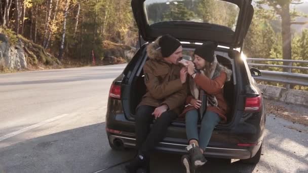 Una pareja amorosa en un viaje por carretera. El tipo y la chica están sentados en el maletero abierto del coche, comiendo sándwiches y alimentándose unos a otros. Parada al aire libre en medio de la carretera. Día de otoño frío y soleado — Vídeos de Stock