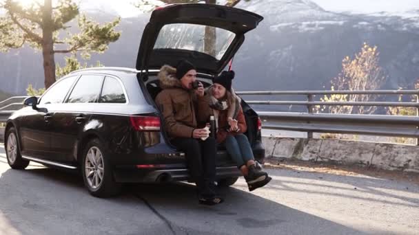 Amante, joven pareja bebe té caliente de termo frasco sentado en el maletero del coche. Feliz pareja caucásica tomando un café durante el viaje por carretera en el campo - beber y comer sándwiches. Admirando el — Vídeos de Stock